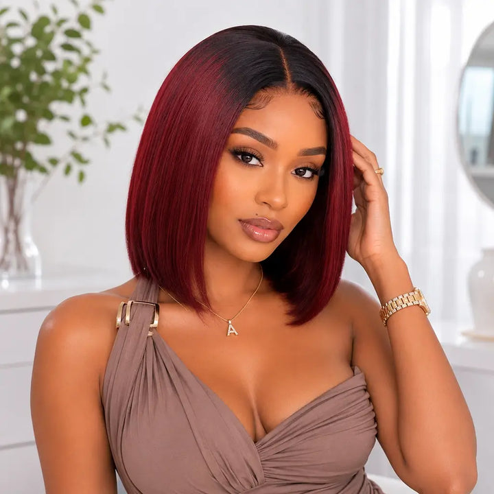 Female model wearing a short straight burgundy bob wig with black roots, elegant summer clothing, gold accessories, and a bright neutral-toned background.